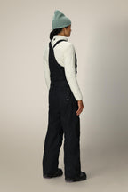 A person stands facing away, wearing a light green beanie, white quilted jacket, black boots, and 686 Black Magic™ 20K Shell Bib women's snow pants by 686, against a plain light background.