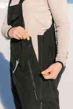 A person is outdoors in bright sunlight, unzipping the side zipper of the 686 Women's Black Magic™ 20K Shell Bib by 686 while wearing a light pink long-sleeve shirt.