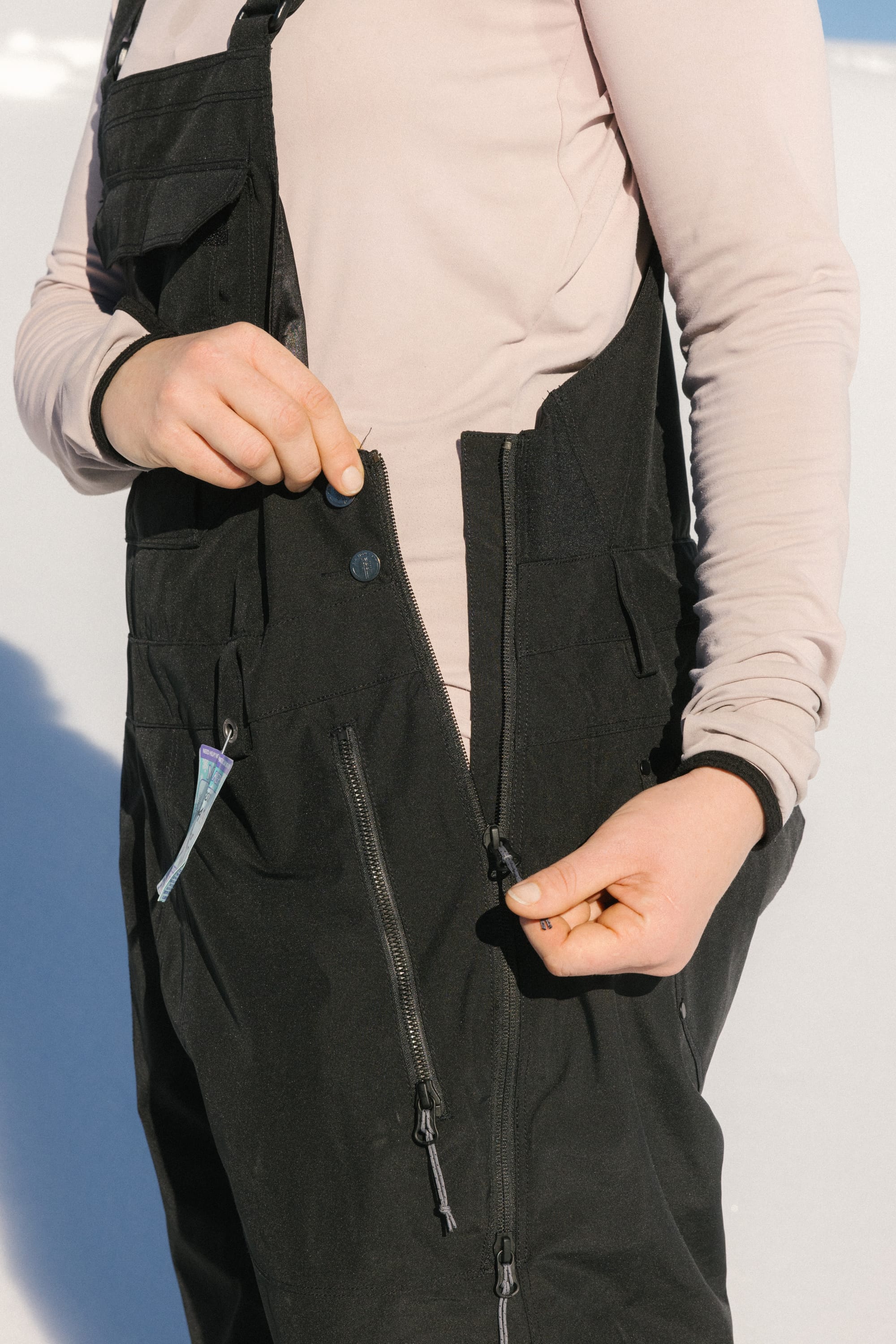 A person is outdoors in bright sunlight, unzipping the side zipper of the 686 Women's Black Magic™ 20K Shell Bib by 686 while wearing a light pink long-sleeve shirt.