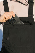 Wearing the 686 Women's Black Magic™ 20K Shell Bib, a woman in a light pink top puts a black smartphone with three camera lenses into its front pocket.