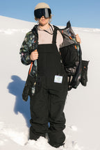 A woman wearing the 686 Women's Black Magic™ 20K Shell Bib stands on snow in a patterned jacket, goggles, and beige beanie, holding her jacket open to reveal the lining beneath a clear blue sky.