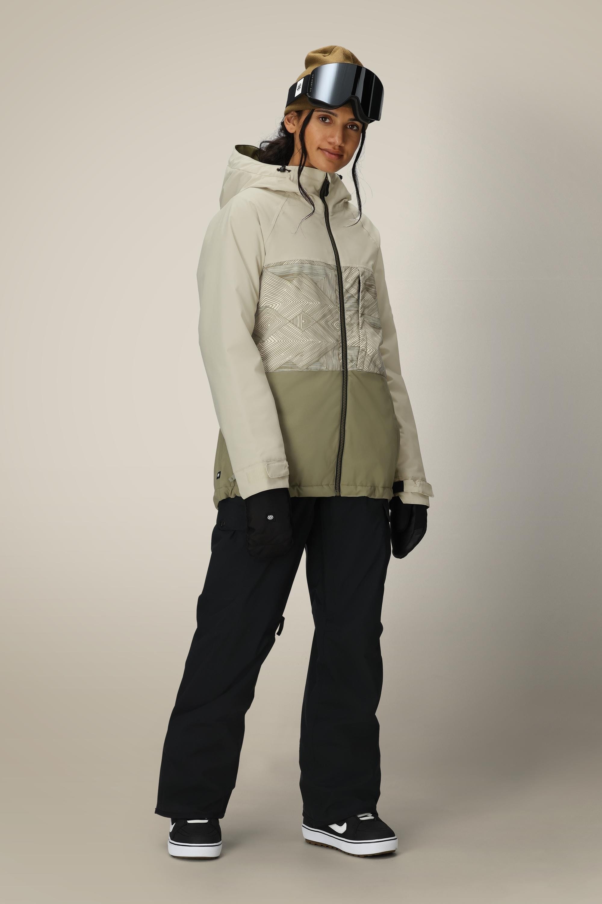 Wearing a light beige and olive green hooded winter jacket, 686 Women's Aura™ Insulated Cargo Pant, gloves, and black snow goggles, a smiling woman stands against a neutral background.