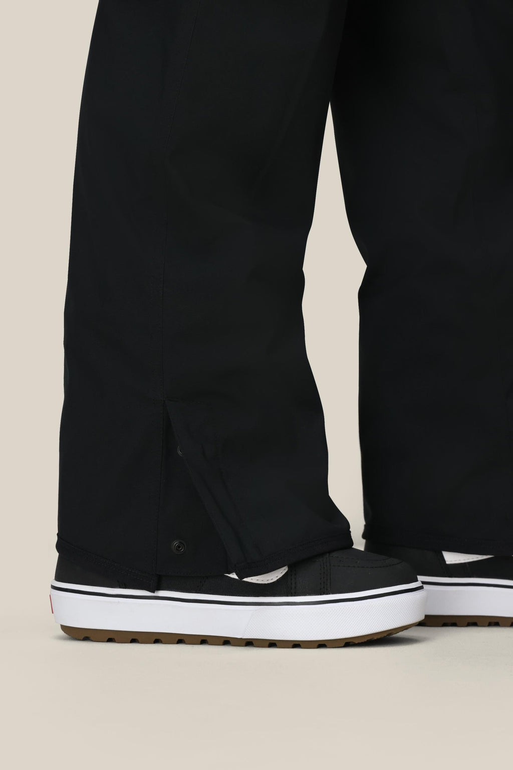 Close-up of a person wearing 686 Women's Aura™ Insulated Cargo Pant with wide legs that cover most of their black and white sneakers with thick, ridged soles, standing on a light beige background.