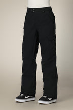 A person wears 686 Women's Aura™ Insulated Cargo Pant by 686—black, waterproof, relaxed-fit with multiple pockets—paired with black-and-white sneakers, standing against a beige background.