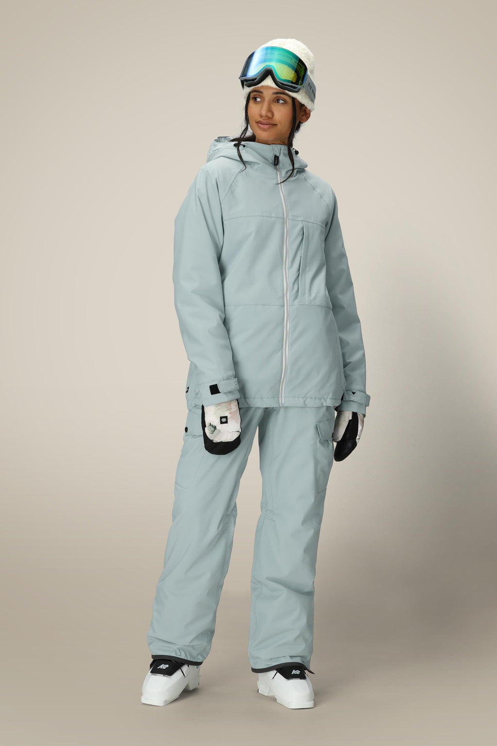 A person wearing a light gray ski jacket, 686 Women's Aura™ Insulated Cargo Pant by 686, white gloves, white ski boots, a white beanie, and ski goggles stands against a plain beige background.