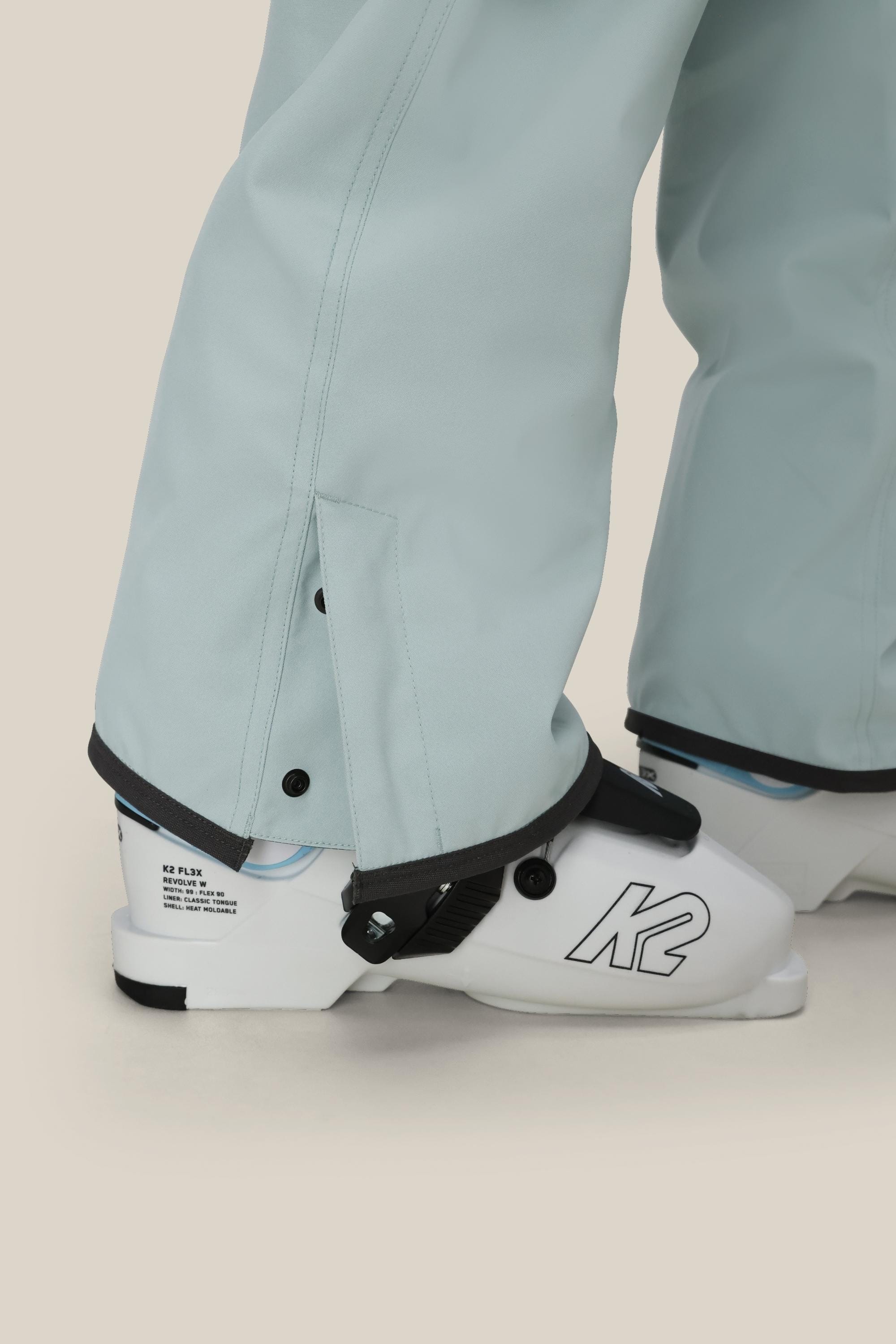 A close-up of a person wearing 686 Women's Aura™ Insulated Cargo Pant in light blue over white K2 ski boots, secured with black fasteners at the ankle. The background is plain beige.