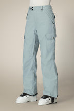 Person wearing 686 Women's Aura™ Insulated Cargo Pant in light blue, featuring waterproof fabric, multiple pockets, and a relaxed fit, paired with white ski boots with K2 branding against a neutral background.