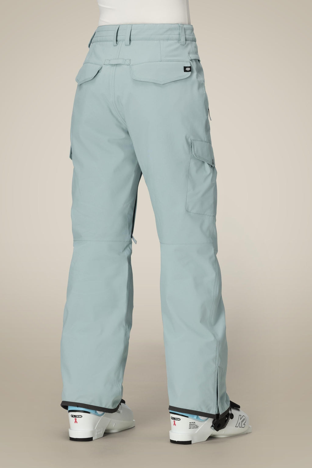 A person wearing light blue 686 Women's Aura™ Insulated Cargo Pants with flap pockets and a relaxed fit, along with white ski boots, stands with their back to the camera against a plain beige background.