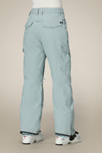 A person wearing light blue 686 Women's Aura™ Insulated Cargo Pants with flap pockets and a relaxed fit, along with white ski boots, stands with their back to the camera against a plain beige background.