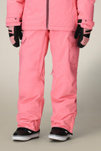 A person wears the 686 Women's Aura™ Insulated Cargo Pant with a pink winter jacket, black gloves with white details, and black-and-white snow boots, standing against a plain background.