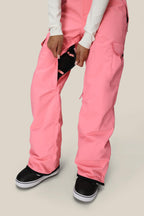 A person wearing bright pink 686 Women's Aura™ Insulated Cargo Pant and black sneakers unzips a thigh vent, revealing black inner fabric. The background is plain and light-colored.