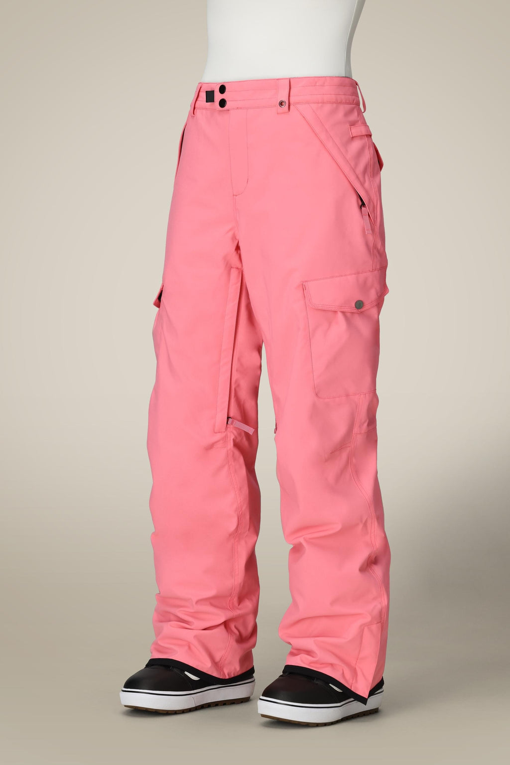 A person models the 686 Women's Aura™ Insulated Cargo Pant in bright pink with cargo pockets and black-and-white snow boots against a beige background. Only the lower body is visible.