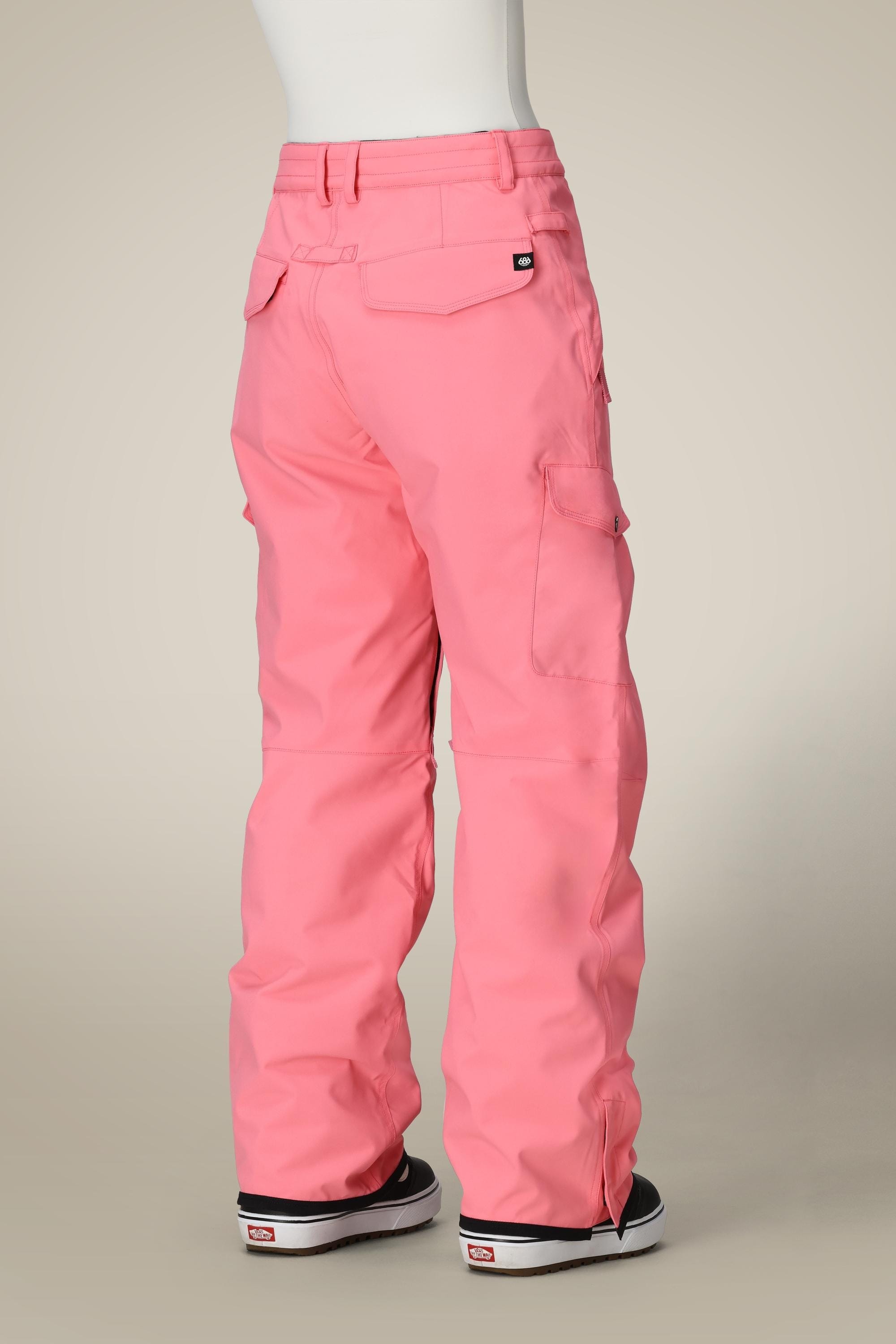 A person stands with their back to a beige background, wearing the 686 Women's Aura™ Insulated Cargo Pant in bright pink and black sneakers with white soles.