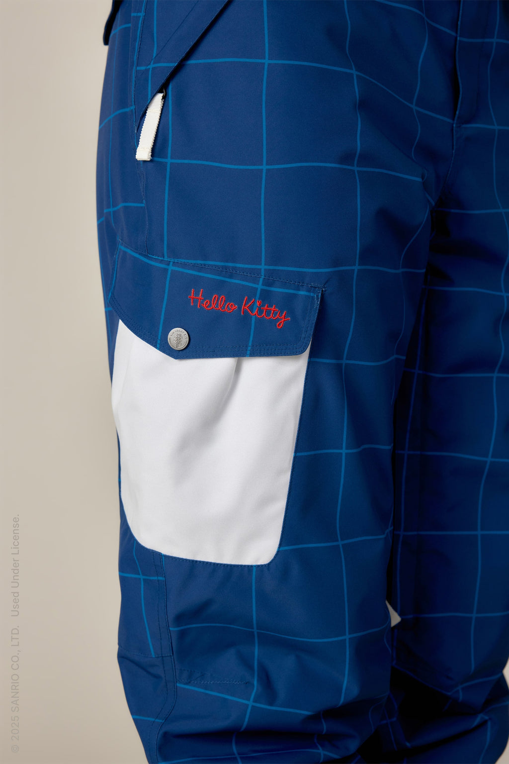 Close-up of 686 Women's Aura™ Insulated Cargo Pant in blue with a white grid pattern, featuring a white pocket flap embroidered with red Hello Kitty text. This modern design by 686 blends functional waterproofing with stylish appeal.