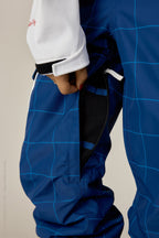 A woman in 686 Women's Aura™ Insulated Cargo Pant by 686, paired with a white jacket, opens a zippered vent on the blue grid-patterned waterproof pants to reveal black mesh lining underneath.