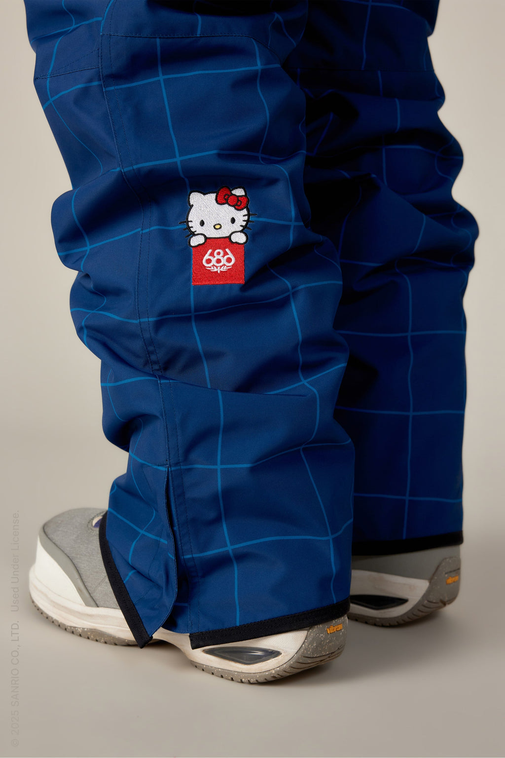 A close-up of someone wearing the 686 Women's Aura™ Insulated Cargo Pant in blue plaid with a small Hello Kitty patch and red 686 logo, paired with gray and white sneakers.