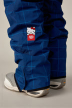 A close-up of someone wearing the 686 Women's Aura™ Insulated Cargo Pant in blue plaid with a small Hello Kitty patch and red 686 logo, paired with gray and white sneakers.