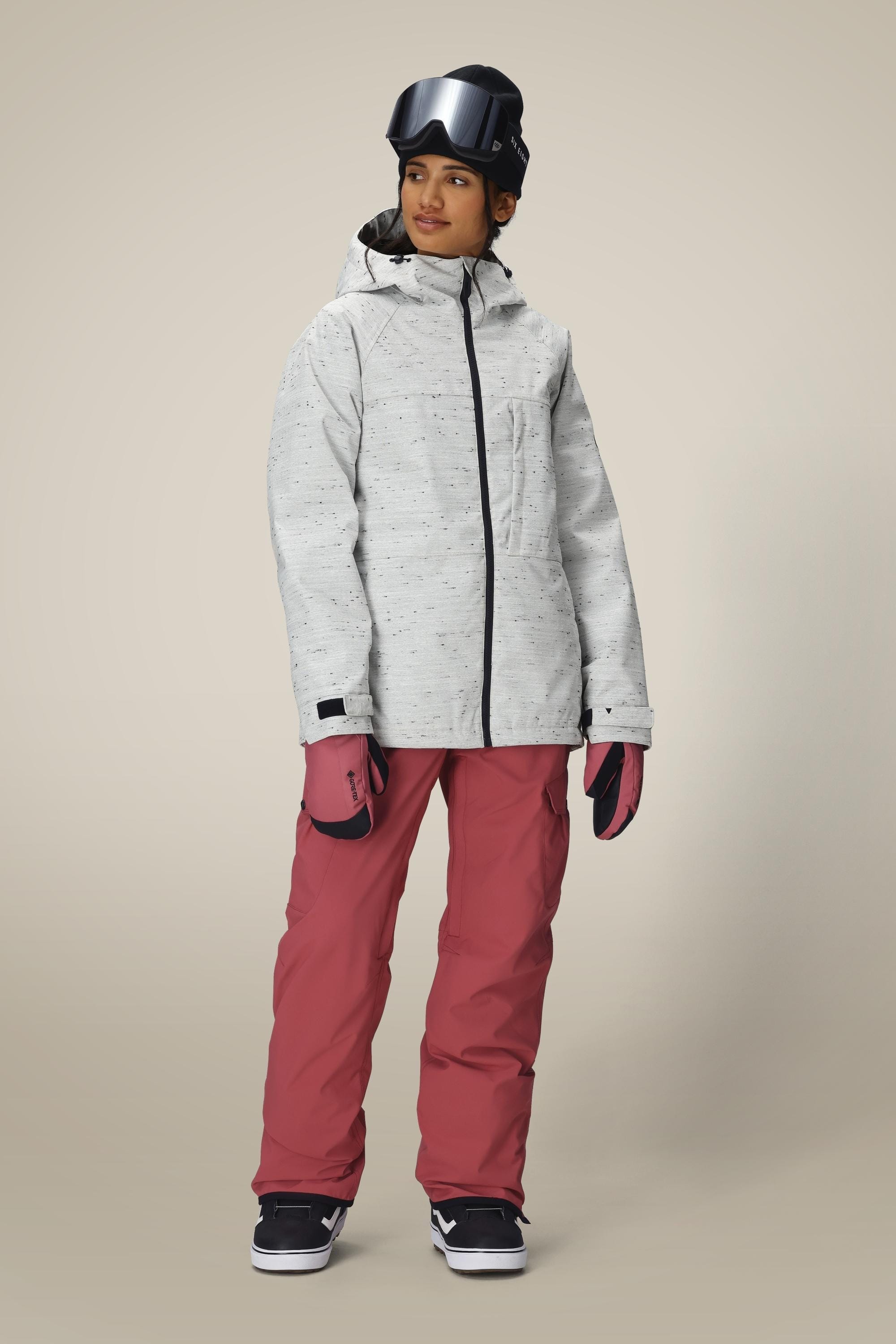 A person wearing a light gray hooded winter jacket, 686 Women's Aura™ Insulated Cargo Pant by 686 in pink, pink gloves, black snow goggles, and black sneakers stands on a neutral background, looking slightly to the side.
