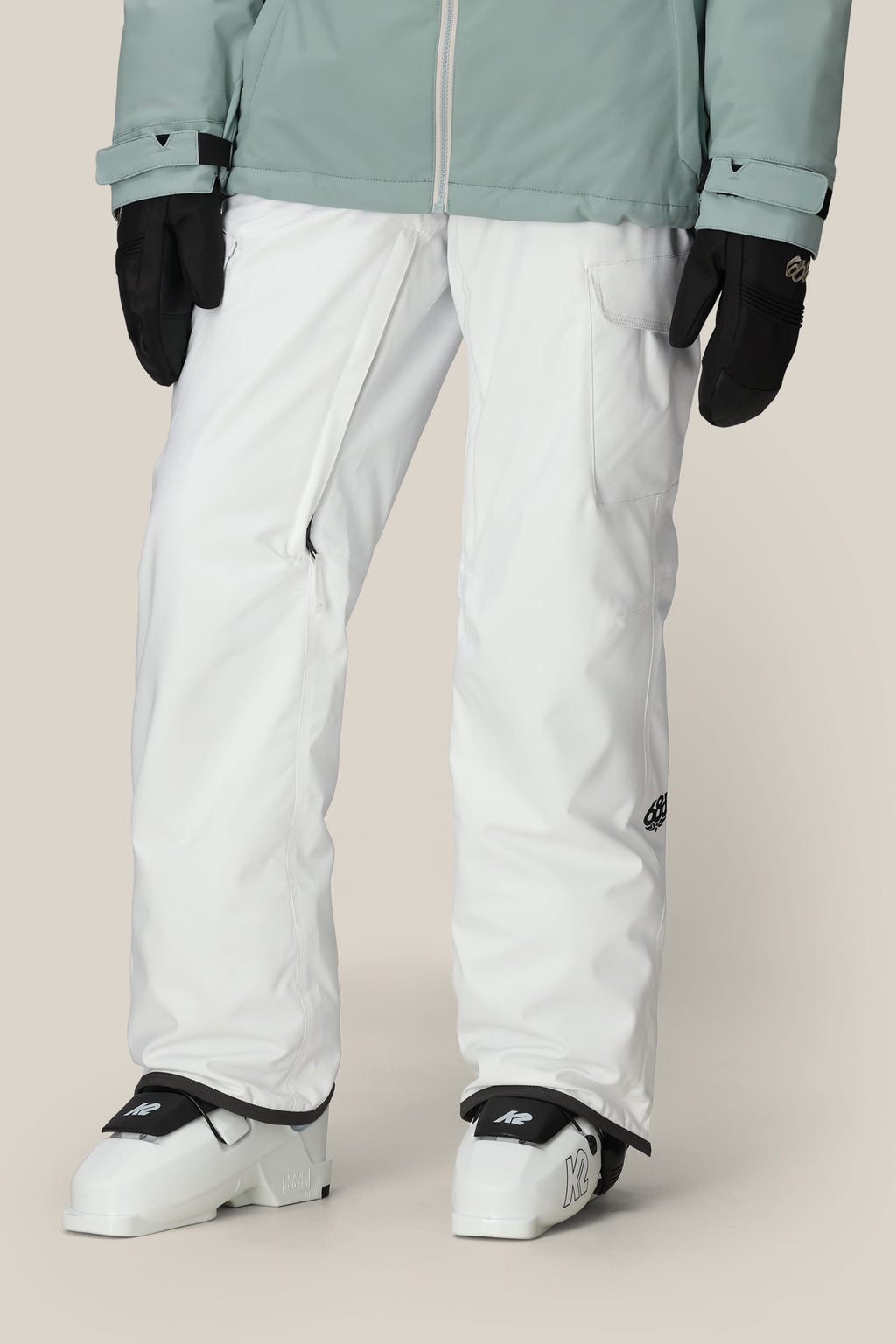 A person wearing 686 Women's Aura™ Insulated Cargo Pant, black gloves, a light green jacket, and white ski boots stands against a neutral background.