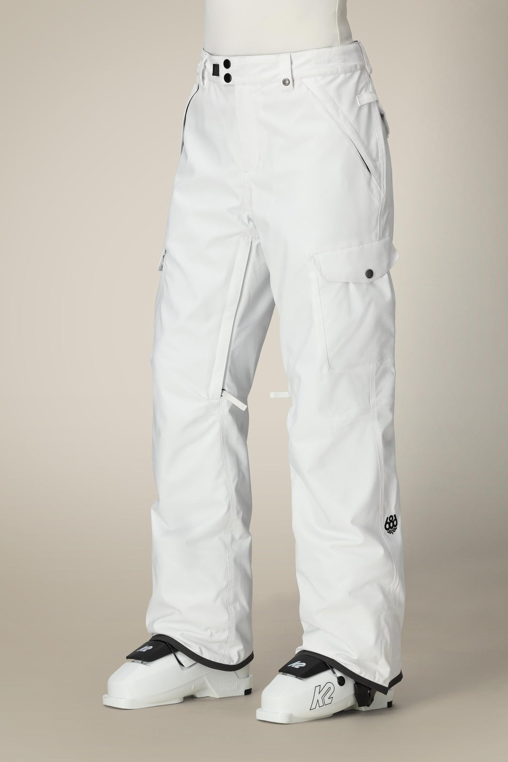 A person wearing 686 Women's Aura™ Insulated Cargo Pant with black buttons and pockets stands in white ski boots labeled K against a plain beige background.