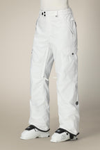 A person wearing 686 Women's Aura™ Insulated Cargo Pant with black buttons and pockets stands in white ski boots labeled K against a plain beige background.