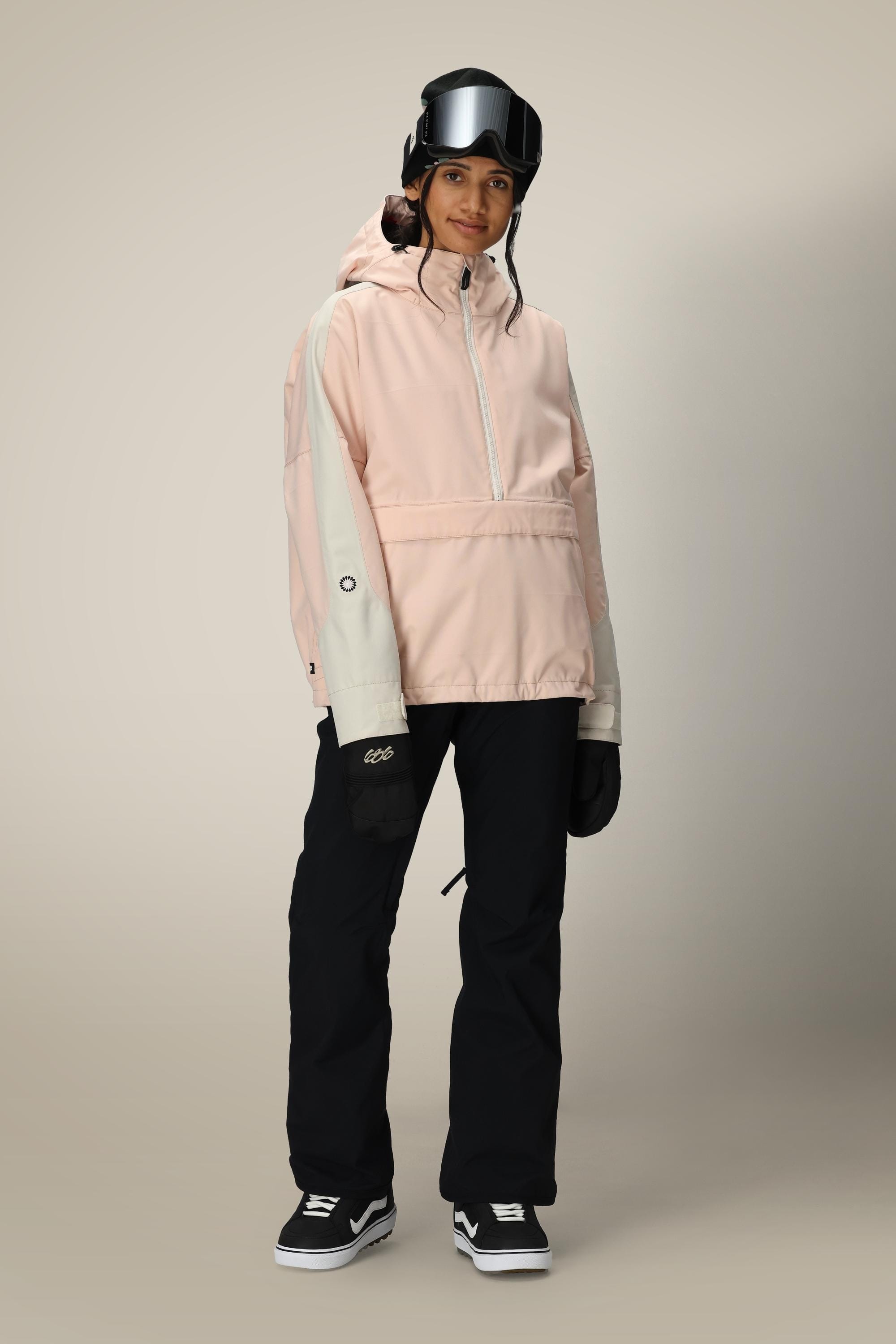 A person wearing a pale pink hooded jacket, 686 Womens Standard Shell Pant in black by 686, black gloves, a black helmet, and ski goggles stands against a neutral background.