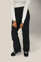A person stands against a neutral background wearing a fitted white long-sleeve top and 686 Womens Standard Shell Pant by 686, holding the pant’s fabric. The look is finished with black and white skate-style sneakers.