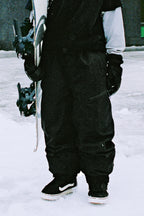 Wearing 686 Women's Outline™ Shell Pant, a person stands on snow holding a snowboard upright. Black gloves and black-and-white boots are also visible, but the person’s upper body and face are not shown.