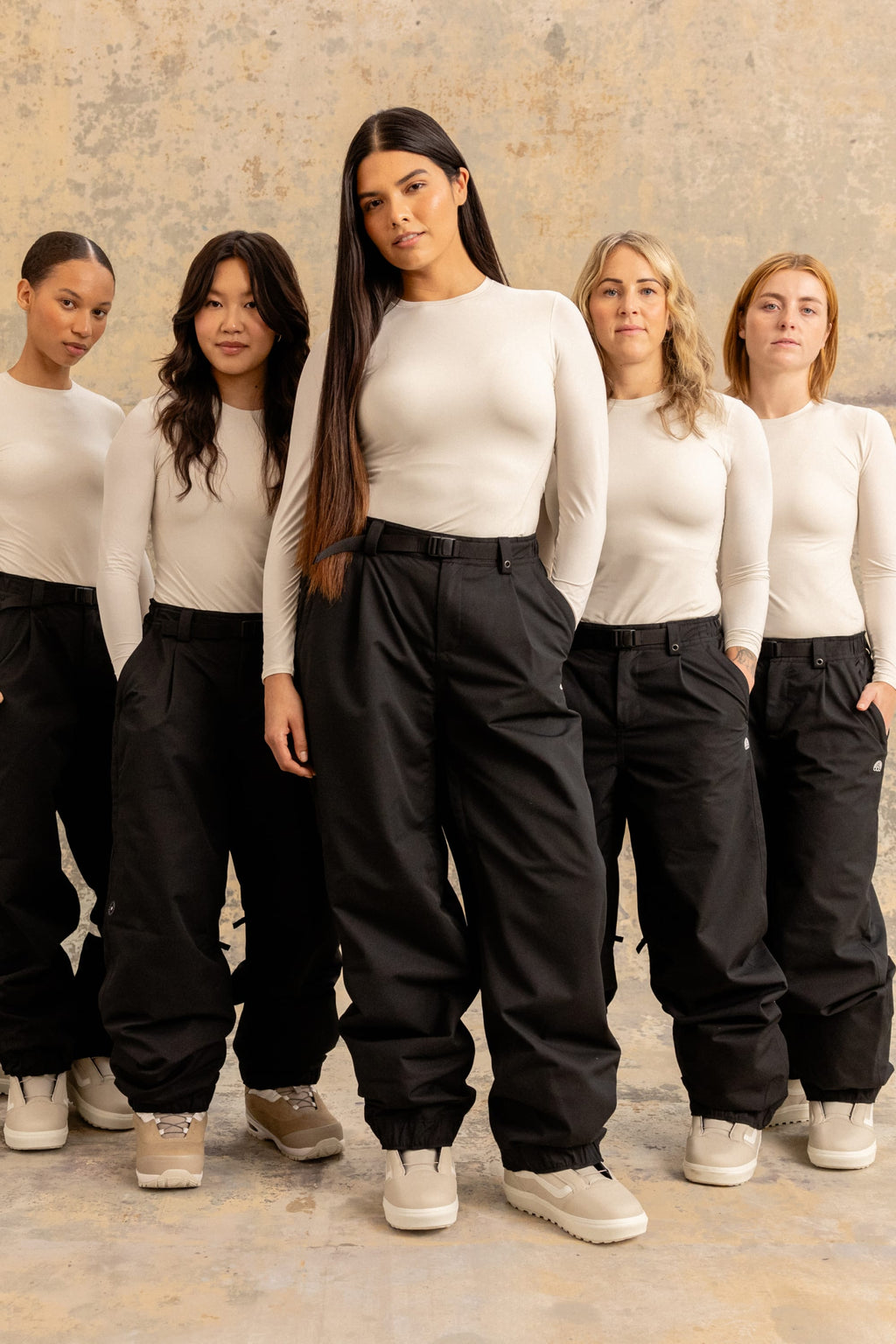 Five women stand together against a textured backdrop, all wearing matching 686 Women's Outline™ Shell Pants with drawstring cuffs and white long-sleeve tops. Their expressions are serious, and they each have different hairstyles.