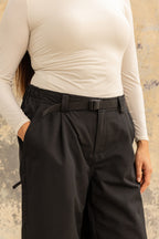 A person wears a fitted, long-sleeve cream top and 686 Women's Outline™ Shell Pant by 686—loose, high-waisted black pants with a belt—standing with one hand in their pocket against a textured beige background.