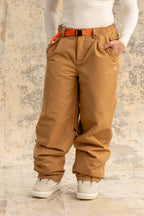 A person stands against a textured beige and gray background wearing 686 Women's Outline™ Shell Pant by 686, styled with an orange belt, white long-sleeve shirt, and white boots.