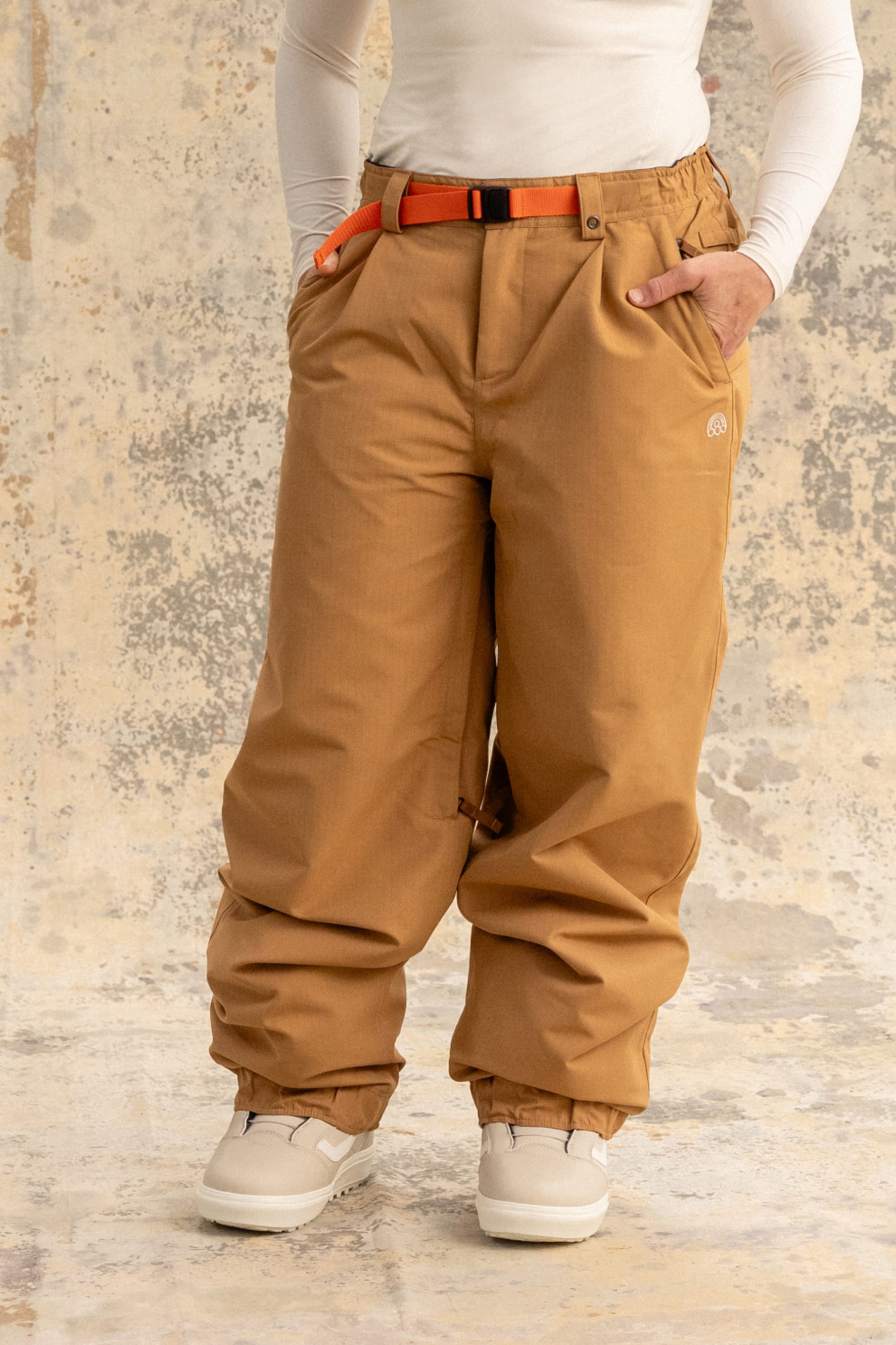 A person stands against a textured beige and gray background wearing 686 Women's Outline™ Shell Pant by 686, styled with an orange belt, white long-sleeve shirt, and white boots.