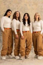 Four women stand side by side in matching 686 Women's Outline™ Shell Pants by 686, paired with white long-sleeve tops and orange belts. They face forward, smiling against a neutral, textured background.