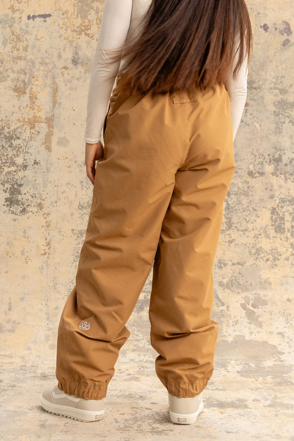 Wearing the 686 Women's Outline™ Shell Pant by 686, a person with long brown hair and a fitted white long-sleeve top stands facing away from the camera against a textured, neutral background.