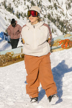 A person wearing 686 Women's Outline™ Shell Pant by 686 stands in the snow with a snowboard. Snowy mountains and another snowboarder are visible in the background.