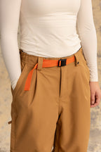 A person wearing a fitted white long-sleeve top tucked into 686 Women's Outline™ Shell Pant by 686, accessorized with an orange belt, stands casually with one hand in their pocket against a neutral background.