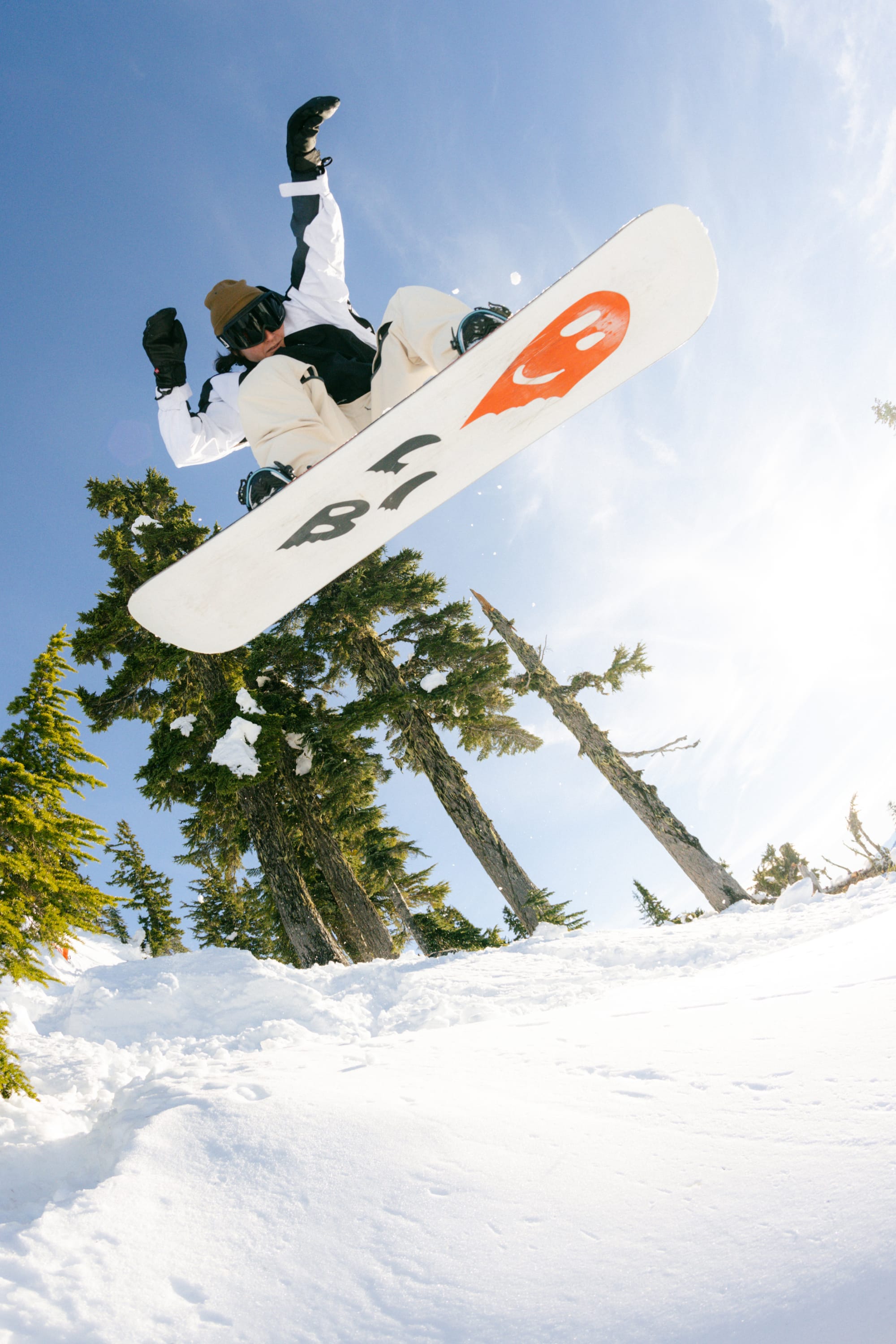 A snowboarder wearing 686 Women's Outline™ Shell Pant and goggles performs a mid-air trick above snowy ground, with pine trees and a clear sky in the background.