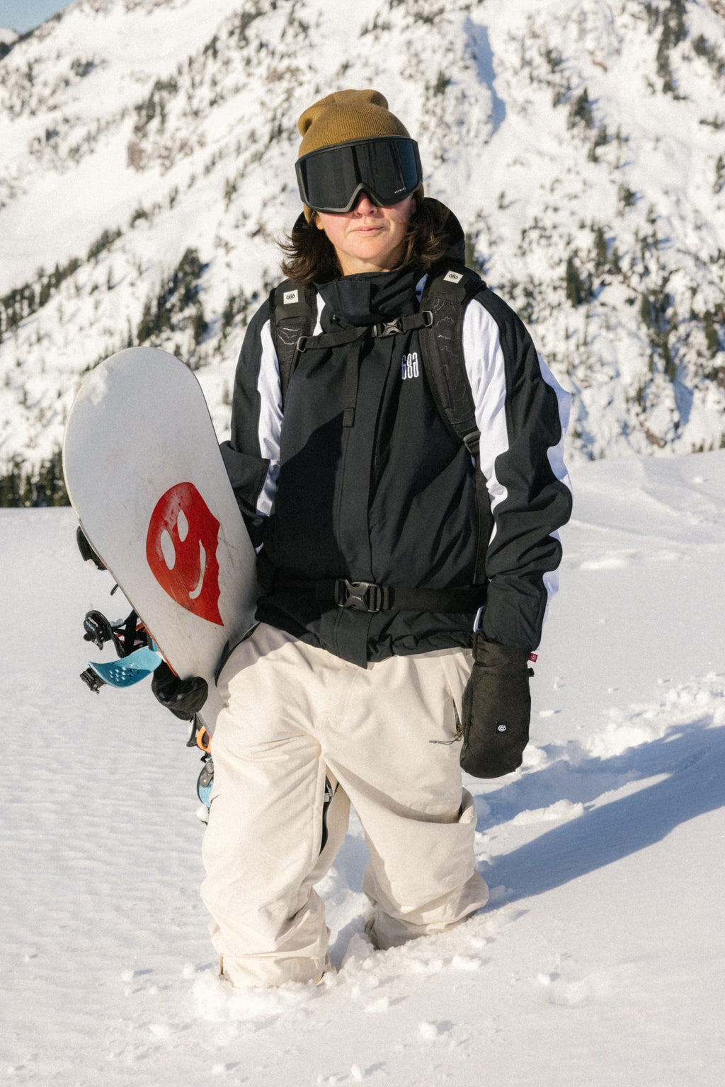 A snowboarder wearing 686 Women's Outline™ Shell Pant by 686 stands in deep snow with a red smiley face snowboard; snow-covered mountains rise in the background.