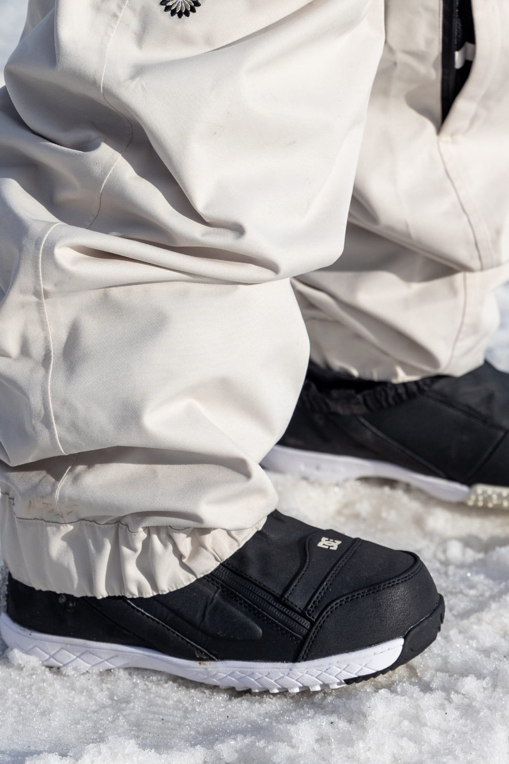 Close-up of a person wearing 686 Women's Outline™ Shell Pant in off-white with black winter shoes, standing on snow.