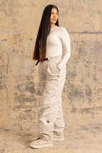 A woman with long straight hair, dressed in a white long-sleeve top, 686 Women's Outline™ Shell Pant by 686 in light beige, and white shoes, stands before a textured beige and gray backdrop, gazing slightly to the side.