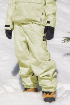A person wearing 686 Womens Sublime Shell Pant in pale green stands on snow, paired with a matching jacket, black gloves, and tan snow boots. Only the lower half of their body is visible.