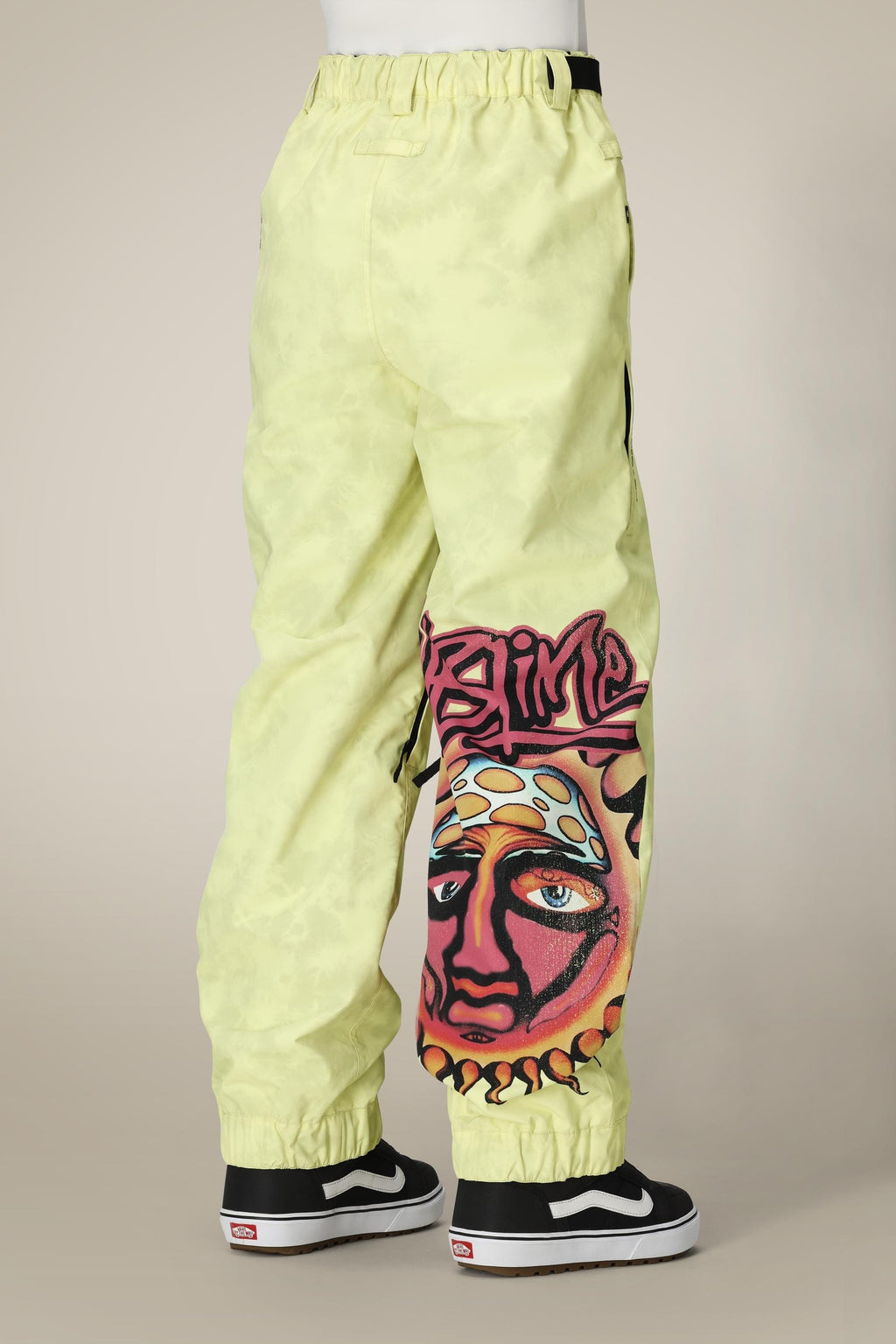 A person wearing 686 Womens Sublime Shell Pant in light yellow with a sun face and SLIME graffiti on one leg, paired with black and white Vans sneakers, shown from the back.