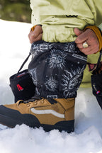 Wearing 686 Womens Sublime Shell Pant, a person in tan and black Vans boots adjusts black snow gaiters with sun and face patterns and Sublime text, standing on snow.