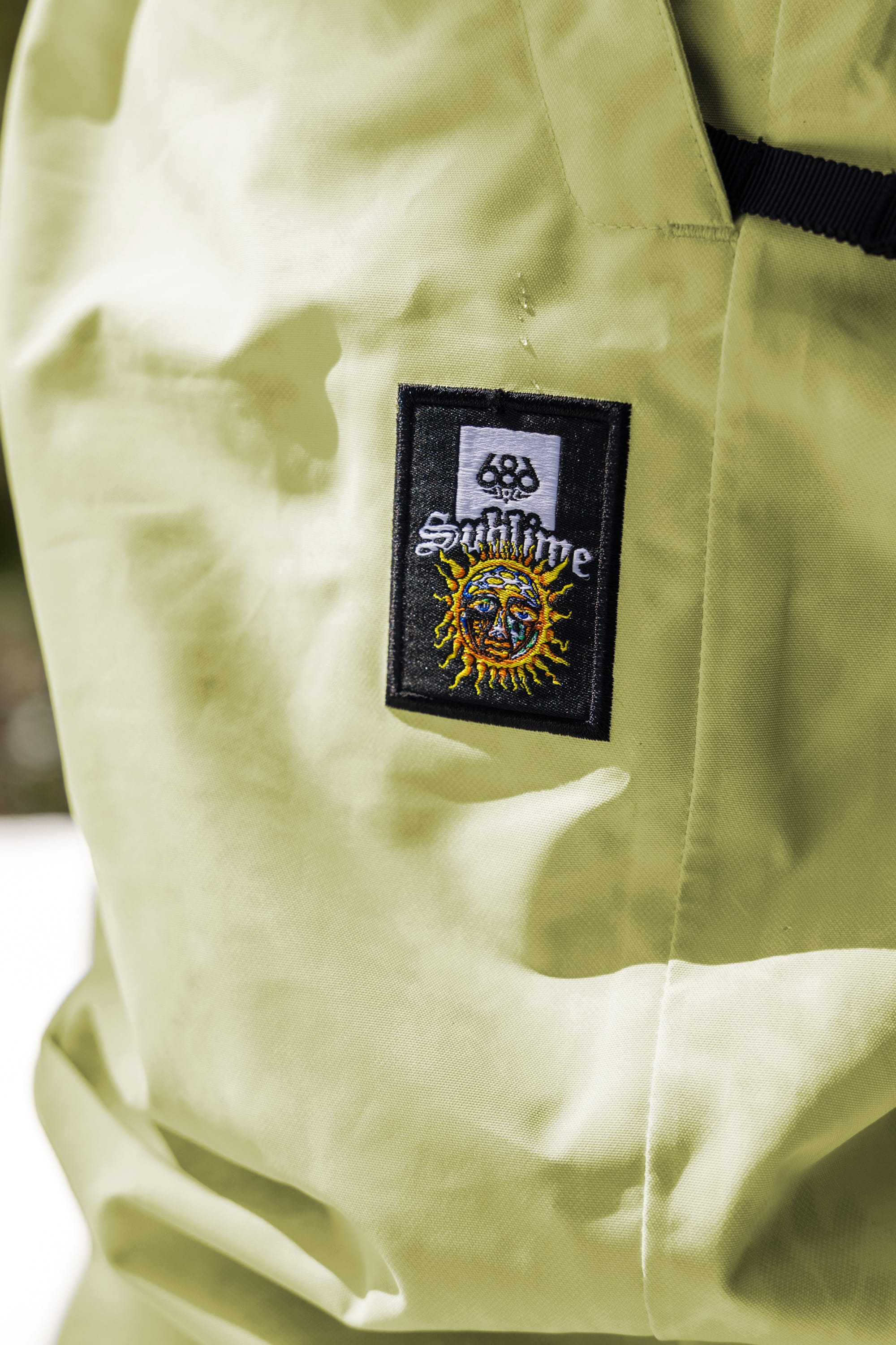 A close-up of a patch on the 686 Womens Sublime Shell Pant, featuring Sublime, a sun-face design, and a smaller logo. Made by 686, the light green waterproof fabric looks both durable and stylish.