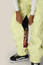 A person wears 686 Womens Sublime Shell Pant in pale yellow with a side zipper partially open, revealing colorful patterned fabric underneath. They pair them with black and white Vans sneakers and a classic white long-sleeve top.