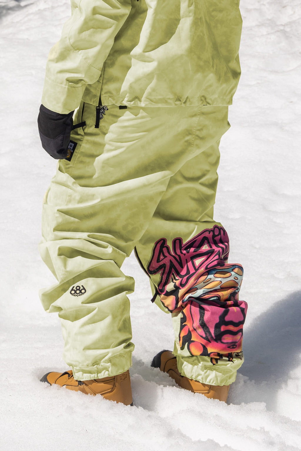 A person stands on snow wearing 686 Womens Sublime Shell Pant in light green, paired with tan boots, black gloves, a matching jacket, and graffiti art knee pads. Only the lower body is visible.