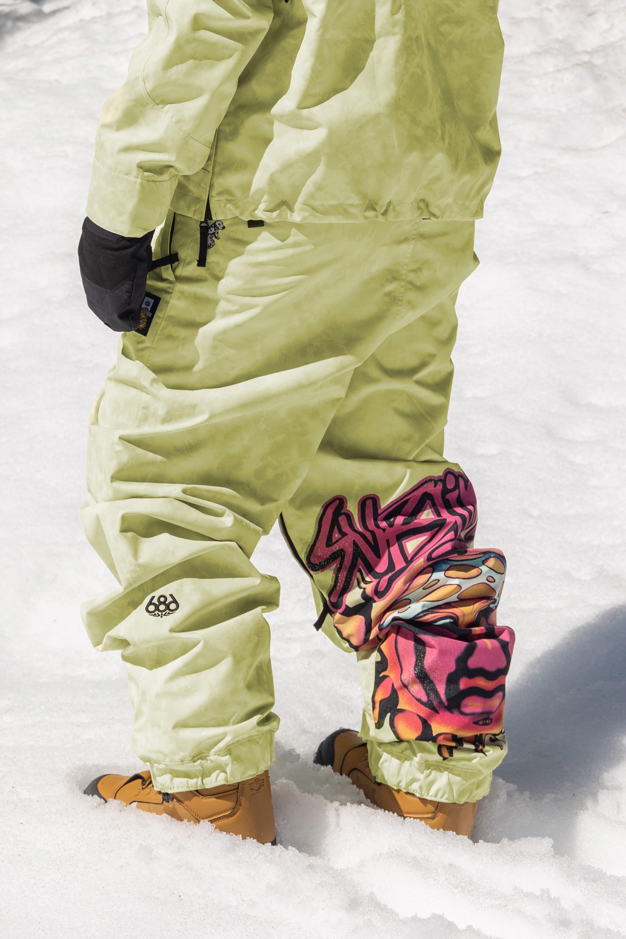 A person stands on snow wearing 686 Womens Sublime Shell Pant in light green, paired with tan boots, black gloves, a matching jacket, and graffiti art knee pads. Only the lower body is visible.