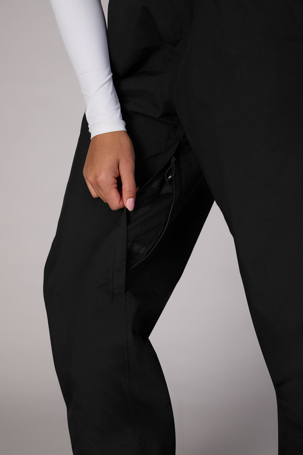 A person opens a zippered pocket on the side of their 686 Women's GORE-TEX Outline™ Shell Pant, wearing a white long-sleeve top. Only their lower torso and hand appear against a plain light background.