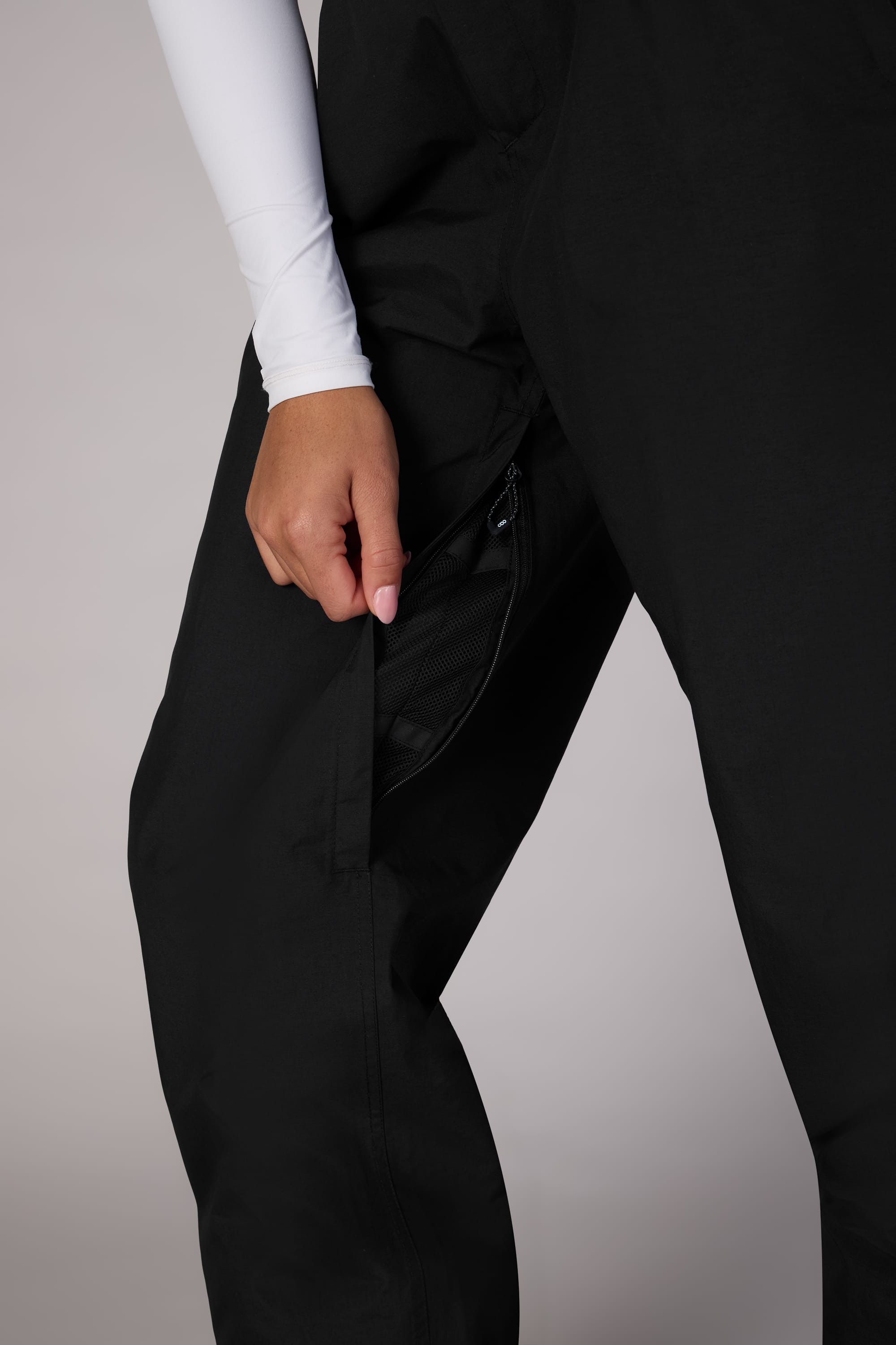 A person opens a zippered pocket on the side of their 686 Women's GORE-TEX Outline™ Shell Pant, wearing a white long-sleeve top. Only their lower torso and hand appear against a plain light background.