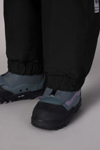 Close-up of a person wearing 686 Women's GORE-TEX Outline™ Shell Pant with elastic cuffs and grey-blue winter boots with black soles, standing on a light-colored surface.
